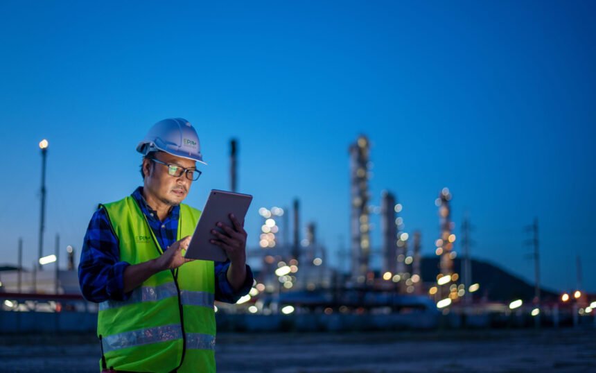 Engineer wearing safety uniform and helmet looking detail tablet on hand with oil refinery factory at night time background.